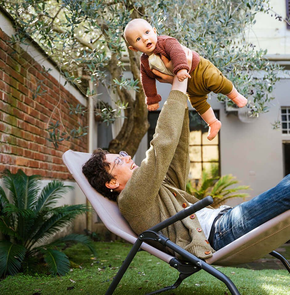 Ein Mann hält ein Baby in die Luft, während er entspannt in einem Stuhl sitzt.